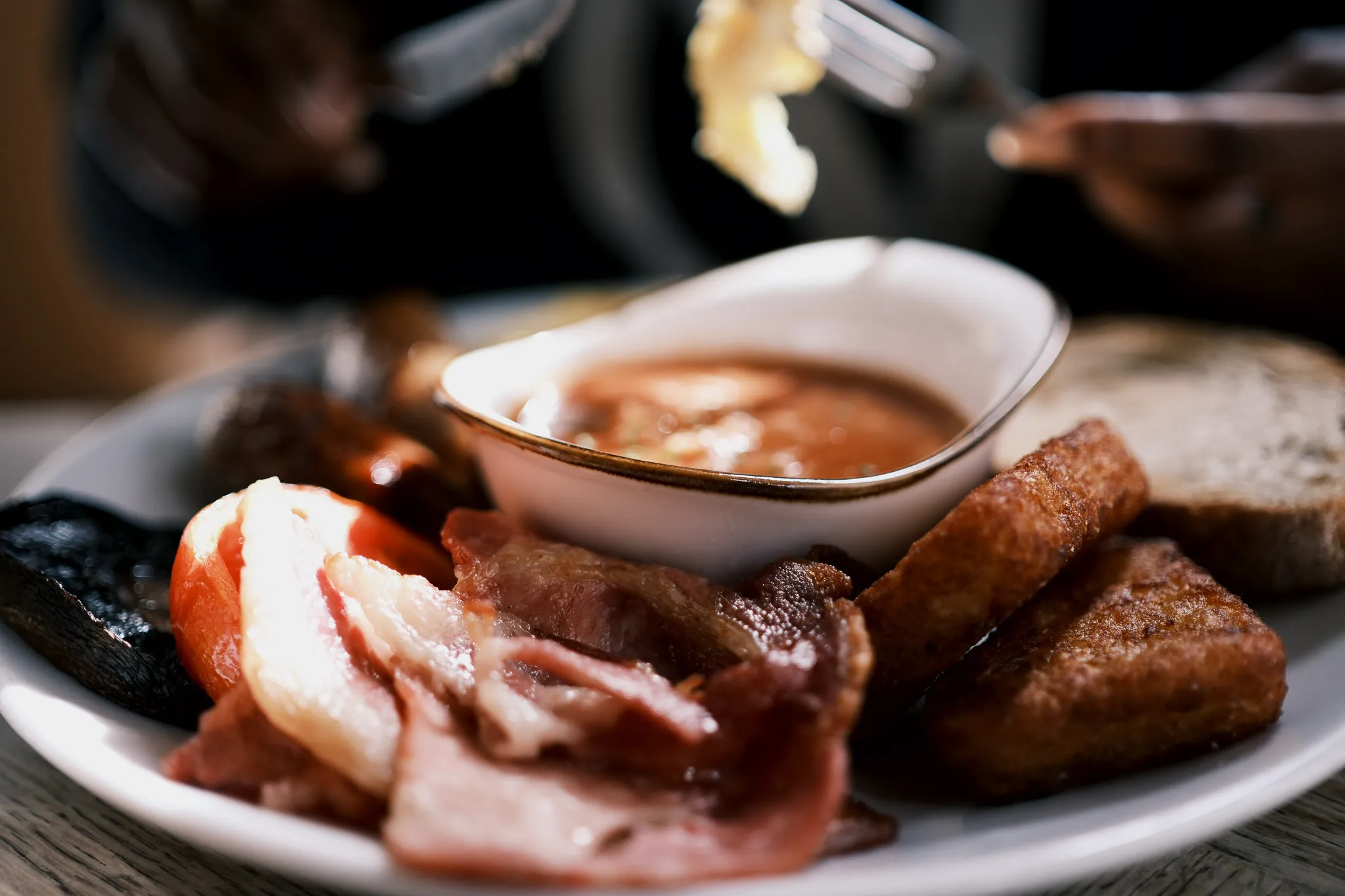 full english breakfast of fluffy eggs, fried bacon, crispy hash browns, mushrooms, tomatoes, thick sausages, toast and a bowl of beans, presented nicely on a plate.